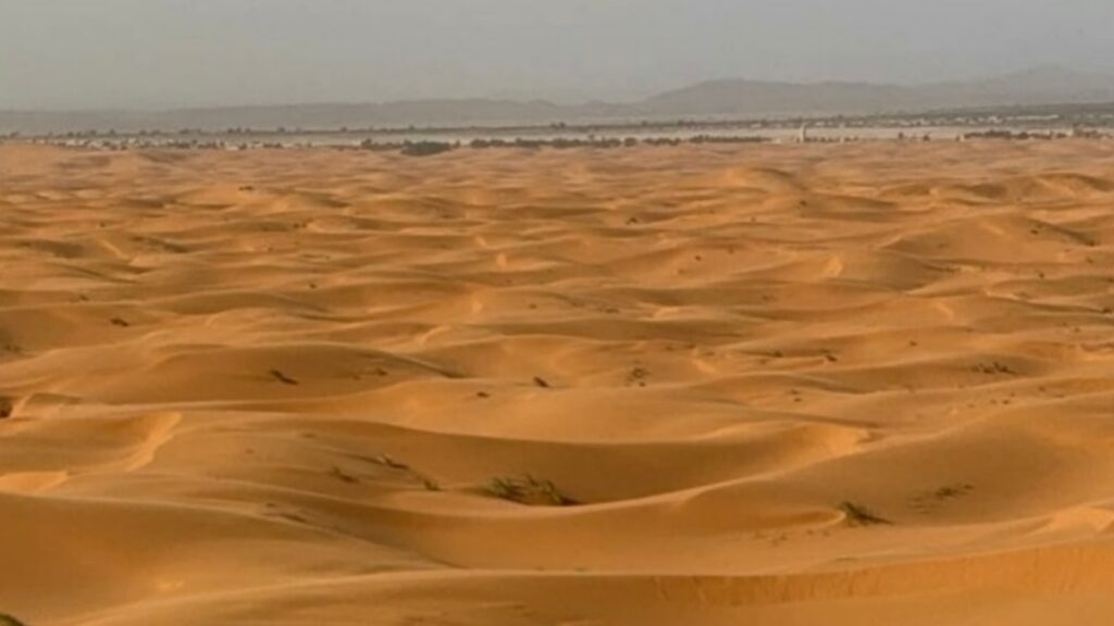 Deportista deslizándose en sandboard por las dunas de Merzouga, amanecer en el desierto del Sahara, campamento bereber al fondo.