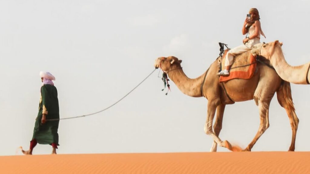 Grupo de viajeros en quad y caravana de camellos en las dunas de Merzouga. Reserva tu tour al desierto de Merzouga con Haya Viajes.