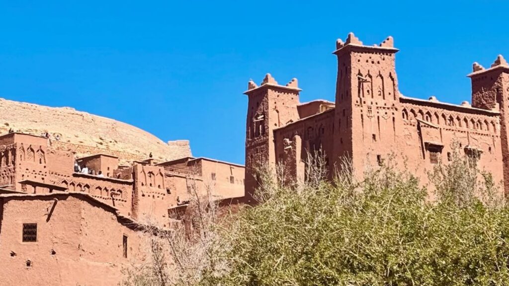 Grupo de viajeros cruzando el río a pie frente a la kasbah de Ait Ben Haddou, con el agua reflejando las murallas de adobe. Reserva tu excursiones desde Marrakech con Haya Viajes.