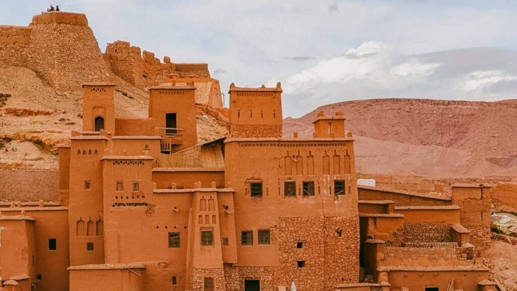 Grupo de viajeros cruzando el río a pie frente a la kasbah de Ait Ben Haddou, con el agua reflejando las murallas de adobe. Reserva tu excursiones desde Marrakech con Haya Viajes.