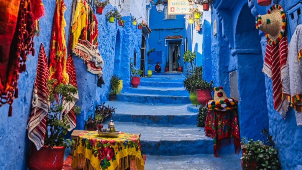 Calles empedradas y fachadas azules en la medina de Chefchaouen, con macetas de flores y un gato descansando. Reserva tu circuito por Marruecos con Haya Viajes.
