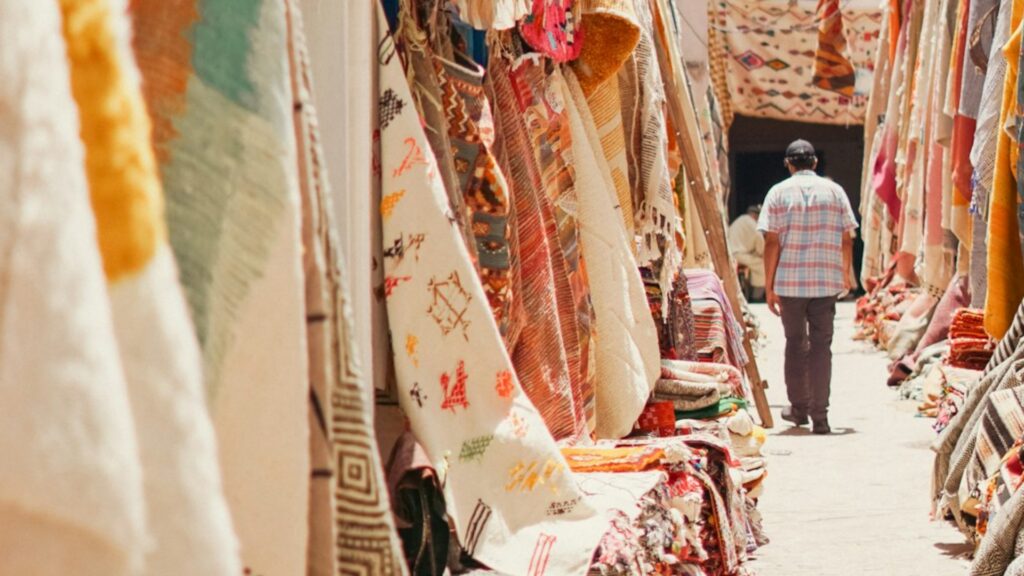 Calle empedrada de la medina de Essaouira con galerías de arte, fachadas blancas y contraventanas azules. Reserva tus excursiones desde Marrakech con Haya Viajes.